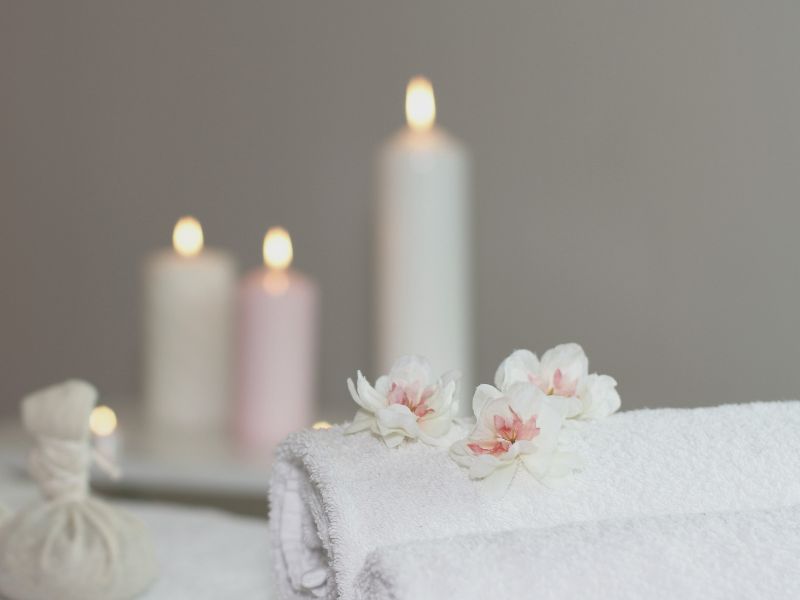 Auf einem zusammengerollten Handtuch liegen Blüten. Im Hintergrund brennen 3 Kerzen. Tara Seymer bietet Ganzheitliche Massagen in Paderborn an.
