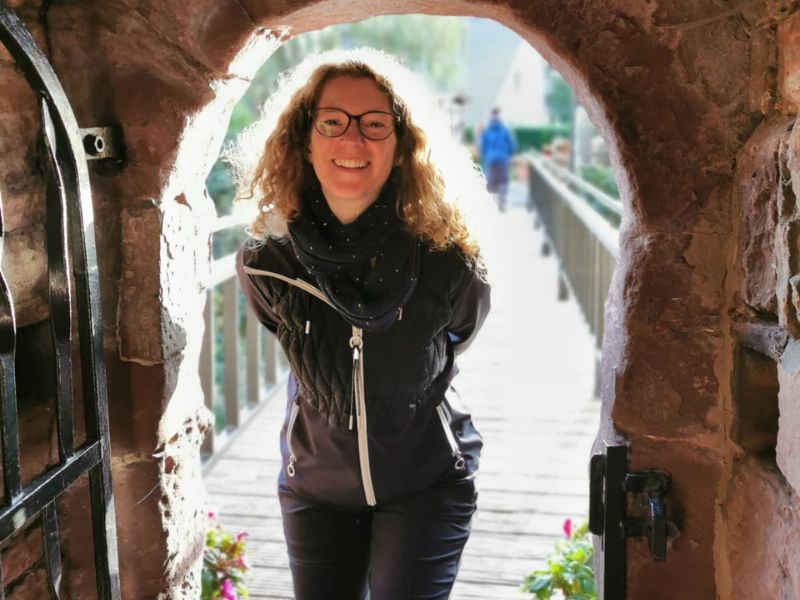 Yogalehrerin Tara Seymer steht lächelnd in einem steinernen Torbogen im warmen Sonnenlicht. Finde passende Yoga Angebote in Paderborn.