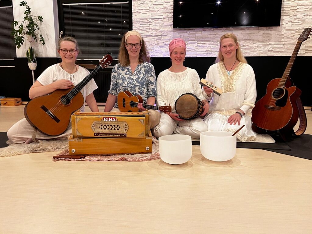 Die Shakti Soulmates Ulrike Menke, Christiane Harbig, Tara Seymer und Tracy Rumpel sitzen entspannt mit Instrumenten wie Gitarre, Ukulele, Trommel und Harmonium zusammen.