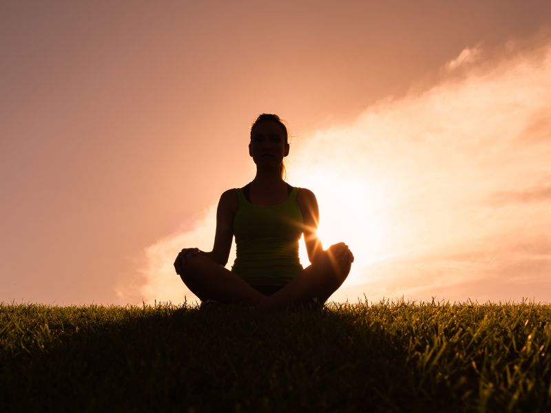 Silhouette einer Frau im Schneidersitz auf einer Wiese im warmen Sonnenuntergang als Symbol, um gelassen Meditation zu lernen.